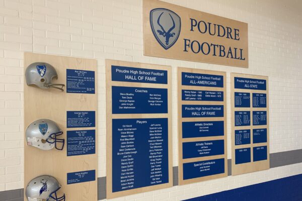 A wall display for Poudre Football featuring two mounted helmets with team logos and several plaques. The plaques list Hall of Fame members, All-Americans, and All-State players, with "Poudre High School Football" prominently displayed at the top.