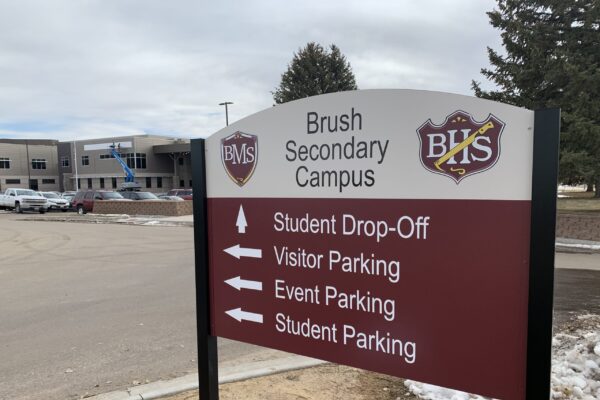 A directional sign at Brush Secondary Campus with arrows indicating locations for Student Drop-Off, Visitor Parking, Event Parking, and Student Parking. The campus building is visible in the background under a cloudy sky.