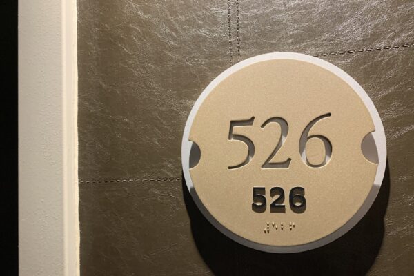 A round beige sign with the number "526" prominently displayed in black numerals is mounted on a gray-tiled wall. The number "526" appears again in smaller print below, with Braille dots below the smaller print.