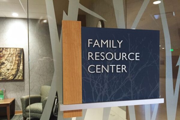A glass door with a wooden and blue sign that reads "Family Resource Center." Inside, blurry furniture is visible, including an armchair and a wall painting. The background has a pattern of frosted stripes or branches on the glass.