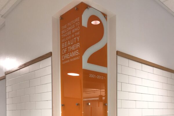 An interior hallway corner featuring a tall orange display with the number "2" and the name "John Bell." The display has a quote about dreams and an illustration of a horse in motion at the bottom. The wall is tiled in white bricks.