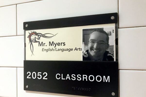 A classroom sign displays "2052 CLASSROOM" at the bottom. Above, there's a smaller sign that reads "Mr. Myers, English/Language Arts" with a profile picture of a man smiling. The sign is attached to a tiled wall.
