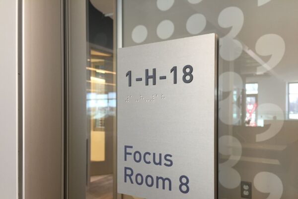 A sign on a glass door reads "1-H-18 Focus Room 8" in black text on a white background. The surrounding area appears to be an office space with a modern interior design, featuring large windows and bright lighting.