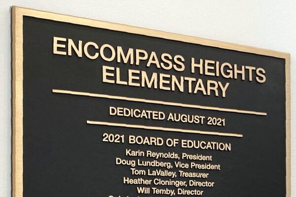 A plaque on a wall entitled "Encompass Heights Elementary," dedicated in August 2021. It lists members of the 2021 and 2017 Boards of Education, the Superintendent, and names the architect and construction company involved in the project.