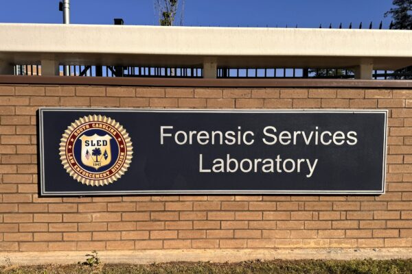 A brown brick wall features a large sign displaying the emblem and text that reads "SLED Forensic Services Laboratory." The sign is rectangular with a black background and white lettering. A patch of grass is visible in front of the wall.