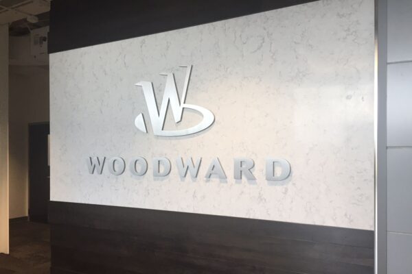 A wall with a sleek, modern design features the embossed logo and name of "Woodward." The logo consists of a stylized "W" above the name "Woodward." The background combines dark and light surfaces, adding contrast to the design.