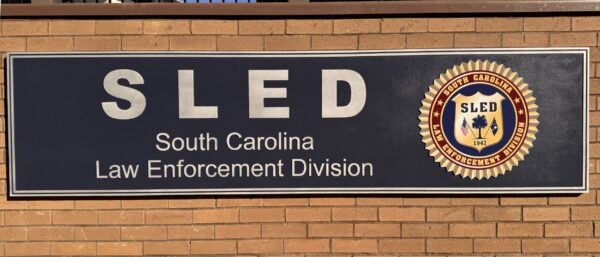 A sign on a brick wall reads "SLED South Carolina Law Enforcement Division" with the SLED shield emblem featuring an eagle and the state seal on the right side.