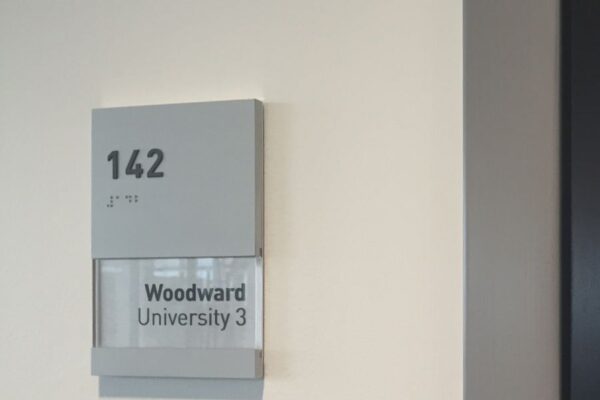 Room sign on a beige wall displays "142" in large numbers at the top left, "2nd Floor" in smaller text below it, and "Woodward University 3" in bold text on a clear panel beneath. The sign is gray with a slightly recessed text area.