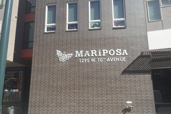 A brick wall with the text "MARIPOSA 1295 W. 10TH AVENUE" in white modern capital letters. To the left of the text is a white butterfly graphic. Shadows and sunlight create a pattern on the wall.