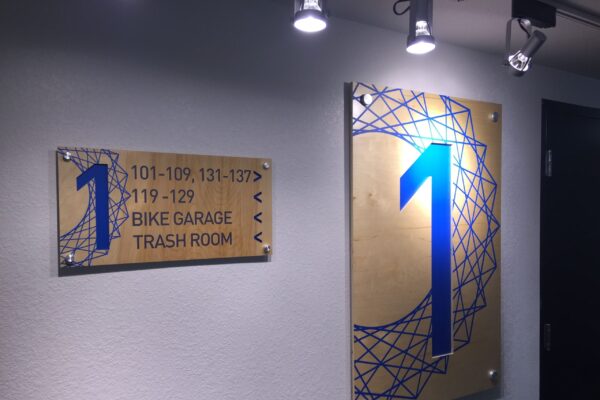 A wall-mounted sign in a hallway displays directions to various apartment units and facilities. The sign has a metallic background and reads, "101-109, 131-137," "119-122," and "Bike Garage, Trash Room" with arrows. Next to it is a large blue number "1.