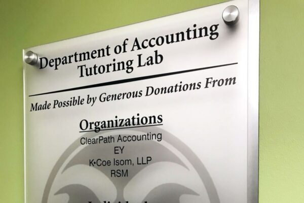 A frosted glass sign in the Department of Accounting Tutoring Lab lists generous donations from various organizations and individuals. The sign includes names like ClearPath Accounting, EY, and several individuals. The background features a subtle university logo.