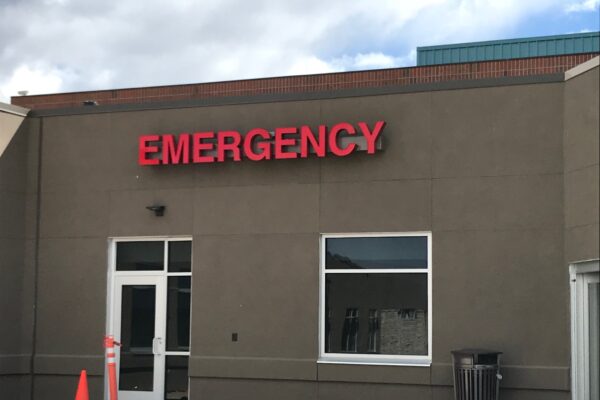 A building exterior with a sign that reads "EMERGENCY" in red letters above a door and window. There are two orange traffic cones placed in front near the entrance. The sky is partly cloudy in the background.