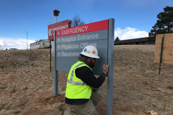 A worker wearing a yellow safety vest and white hard hat installs an outdoor directional sign on a grassy area. The sign indicates the direction for the emergency entrance, hospital entrance, physician offices, and receiving area.