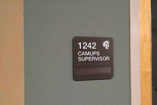 A wall-mounted black sign with white text that reads "1242 CAMUPS SUPERVISOR." There is also an icon of a head wearing a helmet above the text. The sign is mounted on a teal-colored wall.