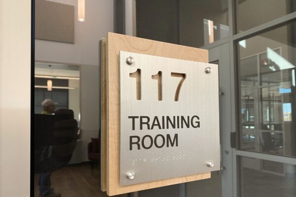 A metallic sign mounted on the wall displays "117 TRAINING ROOM" in bold letters. The sign is attached to a wooden base with screws at each corner. The background shows glass walls and a person seated inside the room.