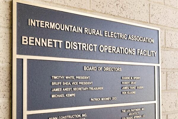 A plaque on a brick wall reads, "Intermountain Rural Electric Association, Bennett District Operations Facility." It lists the Board of Directors, the construction company, and the year 2020. The plaque is bronze with raised lettering.