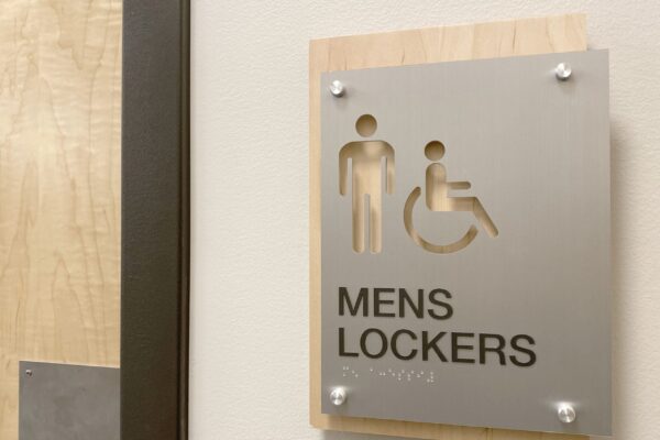 A sign on a wall indicates a men's locker room with accessible facilities. The sign features icons for a man and a person in a wheelchair above the text "Mens Lockers." The sign is mounted on a wooden panel next to a door.