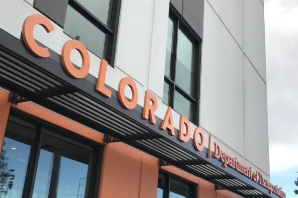 The image shows a building with a sign that reads "COLORADO Department of Revenue" in large, bold letters. The façade has a modern design with large windows and orange accents. The sky is partly cloudy in the background.