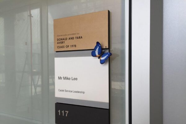 A nameplate on a wall includes layers of different materials. The top plaque reads, "EDWARD AND TAMMY AVERY CLASS OF 1973." The middle plaque reads, "Mr. Mike Lee, District Director Leadership." Below them, there is a room number "117." A blue butterfly decoration is attached.