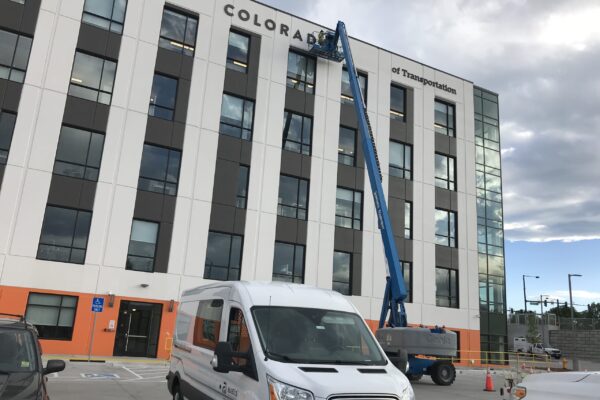 A white van is parked in front of a multi-story building with "Colorado" partially visible at the top. A blue construction lift is extended toward the building's higher floors. The building has large windows and a modern design. The sky is cloudy.