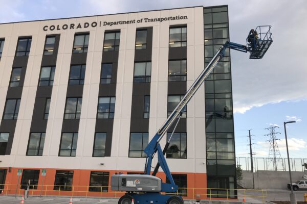 A multi-story building with "Colorado Department of Transportation" in bold letters at the top. A blue construction lift is extended toward the building's higher floors. The building has large windows and a modern design. The sky is cloudy.