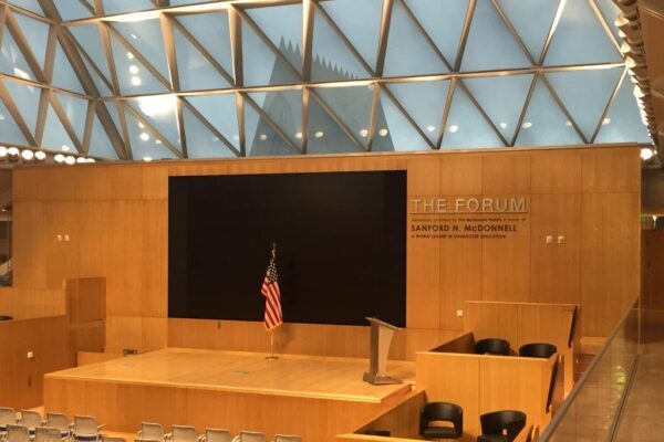 A modern, geometrically designed auditorium named "The Forum" with a large, blank screen flanked by wood paneling. An American flag stands beside a lectern on the stage. Rows of empty chairs and two smaller seating sections are visible.