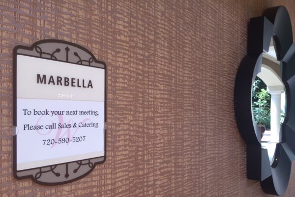 A sign on a patterned wall labeled "Marbella" with contact information for booking a meeting, instructing to call Sales & Catering at 720-904-5207. Next to the sign is a decorative, geometrically-shaped mirror reflecting some greenery.
