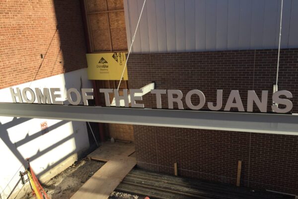 A steel beam with the words "Home of the Trojans" is suspended in mid-air by a construction crane. The background includes a partially constructed building with exposed brick and plywood. The ground below is a mix of dirt and construction materials.