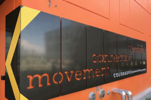 A large, rectangular black sign with yellow accents is affixed to an orange wall. The sign features the words "movement," "connection," and "Colorado" in orange and white text, partially obscured by reflections on the sign's surface.