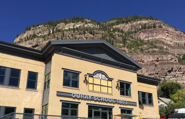 A yellow and gray building labeled "Ouray School 1883" with large windows and an arched roofline stands in front of a backdrop of a rocky, forested mountain under a clear blue sky.