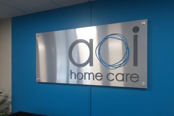 A blue and silver sign mounted on a bright blue wall reads "aoi home care" with a stylized logo featuring three interlocking circles.