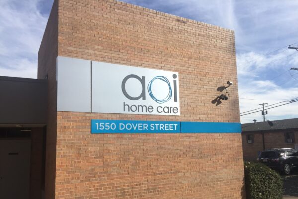 A brick building with a sign that reads "aoi home care" and a blue panel below it displaying the address "1550 Dover Street."
