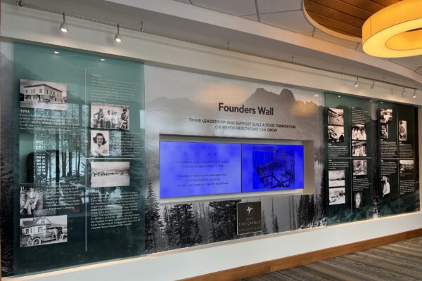 An indoor display titled "Founders Wall" with historical photos and text about the origins of a certain institution or place. The wall has a glass backdrop featuring a mountainous landscape and a central blue-lit screen with additional information.