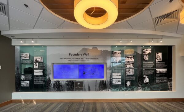 A wall display titled "Founders Wall" showcases black and white photos and informational panels, with a central blue-lit section. Round ceiling lights hang above, and the wall features various images and text commemorating the organization's founders.