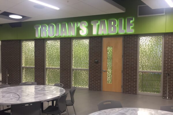 A dining area features a sign reading "TROJAN'S TABLE" in bold, green letters above frosted glass windows and a wooden door. The room includes round tables and chairs with a gray marble-like finish and modern design.