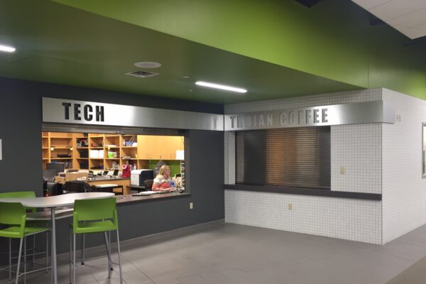 A tech support office next to a closed coffee shop inside a building. The office has a sign that reads "TECH" above a window where two people are working. The coffee shop has a sign that reads "TROJAN COFFEE" above a closed service window. Two green chairs and a table are in the foreground.