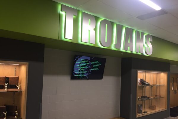 A hallway with a large illuminated sign reading "TROJANS" in green and white. Below the sign is a digital screen mounted on the wall. On either side of the hallway are display cases containing various trophies and awards. A wooden bench is positioned below the screen.