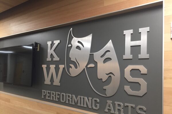 A dark wall with silver metal letters "KWHS Performing Arts" arranged around two theatre masks—one smiling and one frowning—symbolizing comedy and tragedy. The wall is framed by light wood paneling.