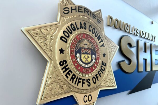 A gold badge with the text "Douglas County Sheriff's Office" and "State of Colorado" is mounted on a wall. The wall also displays large, metallic letters spelling "Sheriff" beside the badge. The badge features intricate design details and the year "1876.