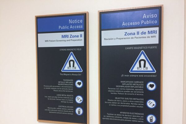 Two informational signs in English and Spanish are displayed on a wall. They provide guidelines and warnings for entering MRI Zone II. Each sign has a blue and black background with white text and includes magnetic field hazard symbols.