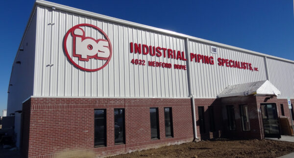 A large industrial building with "IPS" logo and text reading "Industrial Piping Specialist, Inc." on the white upper facade above red brick lower walls. The building is located at "4025 Menoah Road" with blue sky in the background.