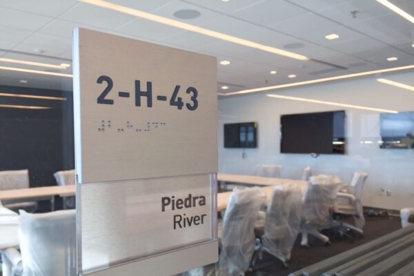 A modern conference room with plastic-covered chairs surrounding a table. The room's glass door has a sign displaying "2-H-43" with Braille text below and "Piedra River" written underneath. Several screens are mounted on the walls inside.