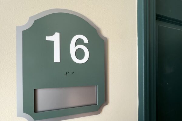 A green and gray door sign with the number 16 prominently displayed in white. Below the number, there is a small line of braille dots. There is a blank rectangular slot near the bottom of the sign. The door next to the sign is slightly ajar.