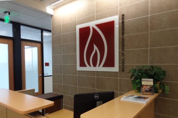 A modern office reception area with a wooden desk, computer screen, and a plant. Behind the desk on the wall is a large red and white logo featuring a stylized flame. Frosted glass doors and a green exit sign are visible in the background.