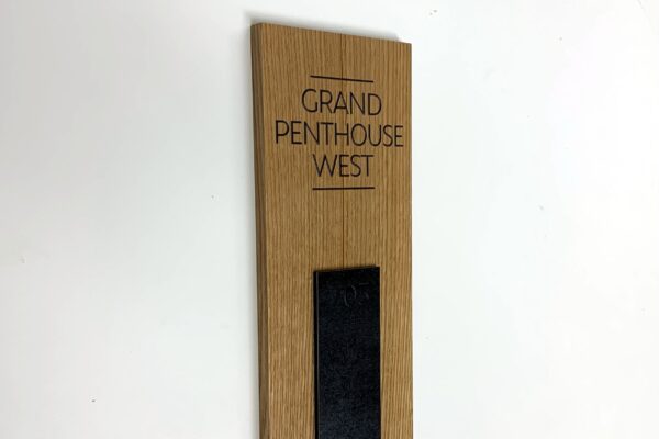 A wooden sign for "Grand Penthouse West" mounted on a white wall, featuring a minimalistic design with black text and a small black vertical strip at the bottom.