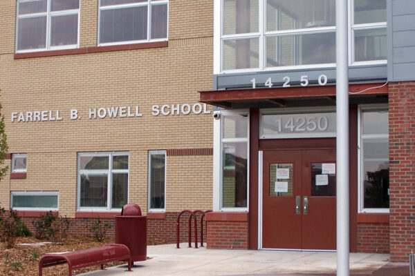 The main entrance of Farrell B. Howell School, displaying the building's name and the address number 14250 above double glass doors. The exterior features red brickwork and beige walls, with windows and a red bench near the entrance.