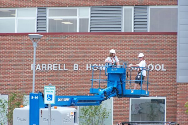 Two construction workers wearing white helmets and safety harnesses work on a blue aerial lift in front of a brick building labeled "Farrell B. Howell School." The building has large windows, and there's a parking sign in the foreground.