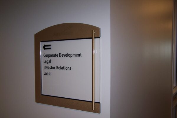 A directory sign on a wall points left with an arrow and lists the departments: Corporate Development, Legal, Investor Relations, and Land. The sign is mounted within a brown wooden frame.
