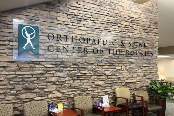 Lobby of the Orthopaedic & Spine Center of the Rockies, featuring a stone wall with the center's logo and name, and a seating area with several chairs and small tables.