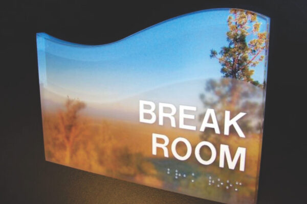 A sign with a scenic background featuring trees and a sky. The sign reads "BREAK ROOM" in large, white letters, and there is Braille text beneath the words. The top edge of the sign has a wave-like curve.